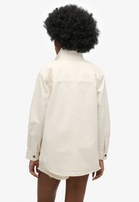 Lys beige oversized skjortejakke lavet af bomuld, med høj krave, knapmanchetter og synlig syning. Bagsiden viser et enkelt design.