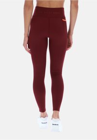 Leggings de tiro alto en color burdeos con tejido texturizado, que cuentan con un acento naranja en el bolsillo y un silueta suave, combinados con zapatillas blancas.