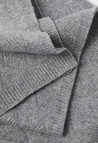 Harmaa kudottu viltti, jossa on herringbone-kuvio. Pehmeä pinta, taiteltu näkyvine saumoineen ja kevyt, tasainen viimeistely.