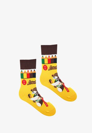 Żółte skarpetki z wzorem brązowego niedźwiedzia trzymającego kanapkę, z brązowym prążkowanym mankietem i kolorowym wzorem ołówków u góry.
