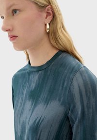 Groene en blauwe tie-dye lange mouwen top met een ronde hals. Model draagt gouden druppeloorbellen. Glad materiaal met subtiele glans.