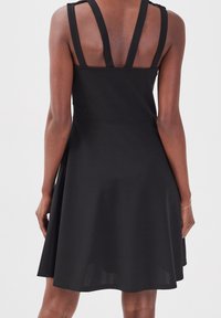 Robe noire sans manches avec des bretelles larges et doubles, portée par une personne vue de dos sur un fond uni.