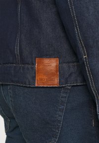 Etichetta in pelle marrone su giacca in denim blu scuro sopra jeans coordinati con cuciture visibili e texture del tessuto.