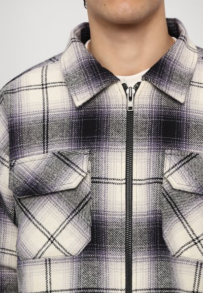 Plaid shirt with a cream, black, and purple color scheme. Features a zip front, collar, and two chest pockets. Made from a textured fabric.