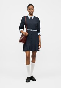 Ženska, oblečena v mornarsko modro nabrano obleko z belim ovratnikom in manšetami, belimi nogavicami do kolen, črnimi mokasini, velikimi očali in rjavo torbo čez ramo.