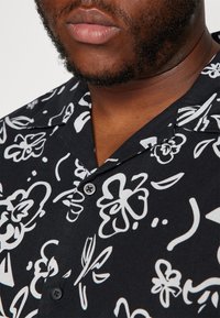 Kortärmad svart skjorta med vita blommiga och abstrakta mönster, med krage och två knappstängningar nära halsringningen.