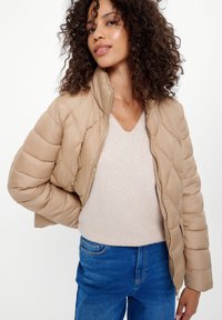 Beżowa pikowana kurtka puchowa z zapięciem na zamek i wysokim kołnierzem, noszona na jasnobeżowym swetrze z dekoltem w serek, w połączeniu z niebieskimi dżinsami.