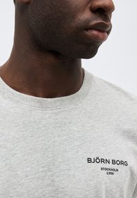 Κοντινό πλάνο ενός ατόμου που φοράει ανοιχτό γκρι T-shirt με την επιγραφή «BJÖRN BORG STOCKHOLM 1956» σε μαύρο χρώμα στο στήθος.