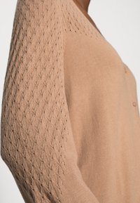 Cárdigan de punto marrón claro con un patrón texturizado en las mangas y un cuerpo liso. Presenta detalles de botones en la parte delantera.