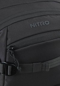 Sac à dos noir en tissu durable, comprenant une poche zippée, une sangle plate et un logo de marque. Texture lisse avec un design minimaliste.