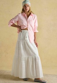 Svetloružová pruhovaná košeľa na gombíky, biela maxi sukňa s očkovými vzormi, vrstvený dizajn a modré ploché topánky. Modelka má na sebe vzorovaný šál.