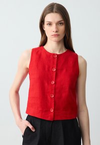 Top senza maniche in lino rosso con scollatura rotonda e sei bottoni frontali. Il modello lo indossa con pantaloni neri a vita alta.