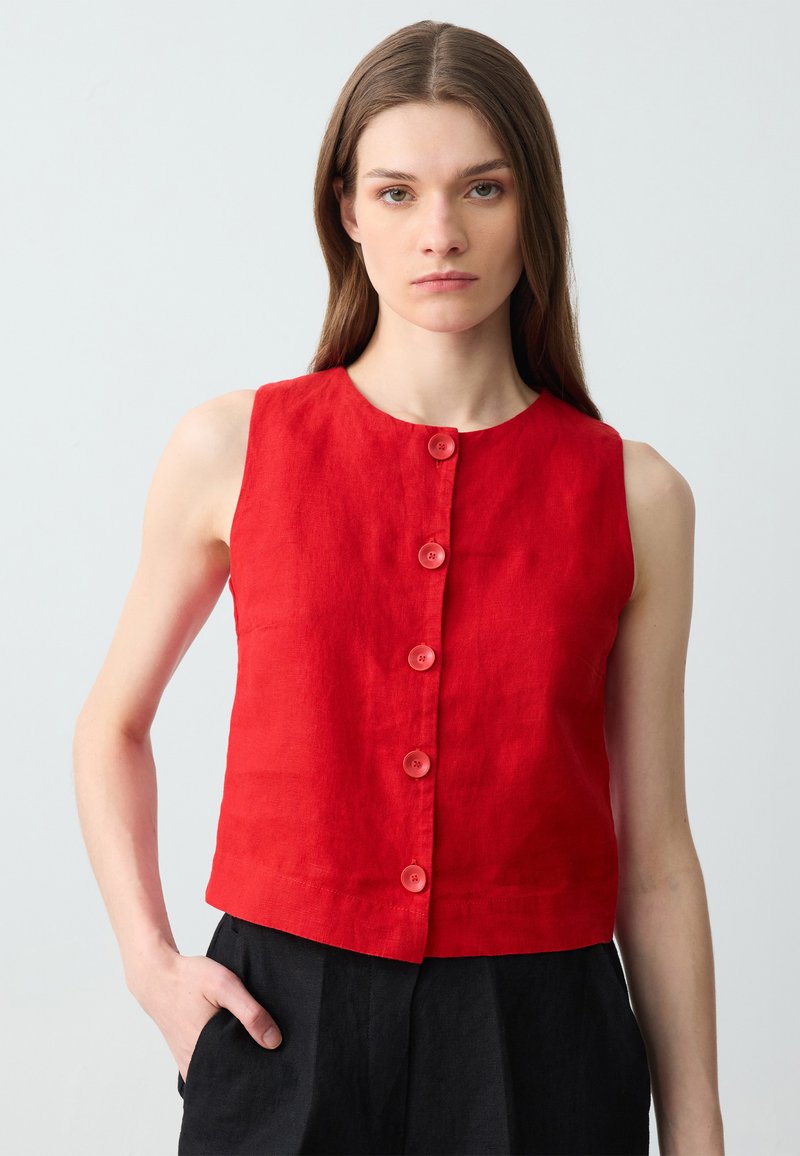 Top senza maniche in lino rosso con scollatura rotonda e sei bottoni frontali. Il modello lo indossa con pantaloni neri a vita alta.