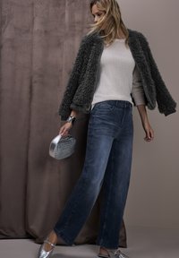 Bomber jacket in pelliccia sintetica grigia sopra un top in maglia chiara, abbinato a jeans a gamba larga blu e ballerine argento, tenendo in mano una borsa metallica intrecciata.