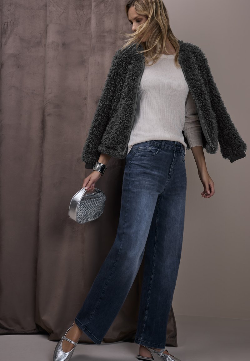 Bomber jacket in pelliccia sintetica grigia sopra un top in maglia chiara, abbinato a jeans a gamba larga blu e ballerine argento, tenendo in mano una borsa metallica intrecciata.