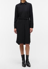 Abito nero con maniche lunghe, chiusura frontale con bottoni e una cintura abbinata. Tessuto liscio con una silhouette sartoriale e orlo al ginocchio.