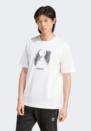 T-shirt bianco in cotone con una grafica di scarpe nere con lacci, centrata sotto il logo "adidas" in grassetto nero. Maniche corte, vestibilità rilassata.