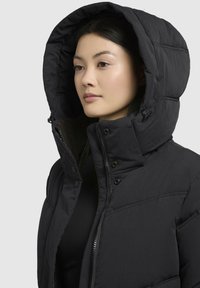 Geacă puf din negru, cu guler înalt și glugă mare, căptușită; prezintă închideri cu fermoar și capse; textură quilted și design elegant.