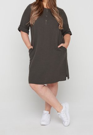 Donkergroene jurk tot op de knie met korte opgerolde mouwen, knoopsluiting, zijzakken en een losse pasvorm. Gecombineerd met witte sneakers.