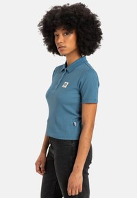 Polo shirt bleu à rayures avec un col et un petit logo brodé sur la poitrine. Il a des manches courtes et une coupe ajustée. Jeans noirs portés en dessous.