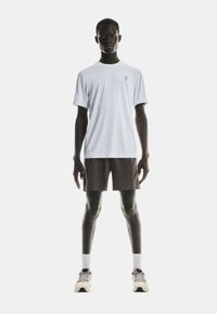 Männlicher Model steht in einem weißen kurzärmligen Sportshirt, dunklen Shorts, weißen Socken und hellen Laufschuhen vor einem schlichten Hintergrund.