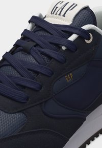 Baskets bleu marine en tissu texturé, avec des lacets épais, une étiquette blanche sur la languette et un logo GAP doré sur le panneau latéral.