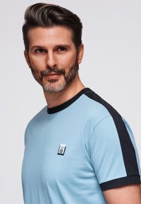 Světlomodré tričko s krátkými rukávy, černými akcenty na ramenou, kulatým výstřihem a malou nášivkou na hrudi s kontrastními barvami.