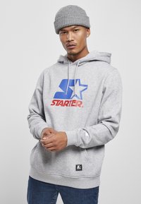 Grå hoodie med et blåt og rødt "STARTER" logo, snore og kængurulomme. Båret med en ribbet grå beanie og denim jeans.