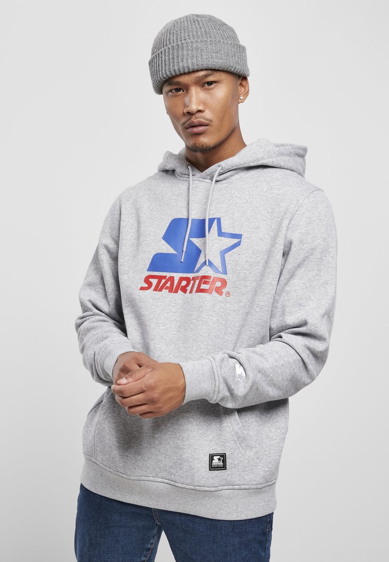 Grå hoodie med et blåt og rødt "STARTER" logo, snore og kængurulomme. Båret med en ribbet grå beanie og denim jeans.