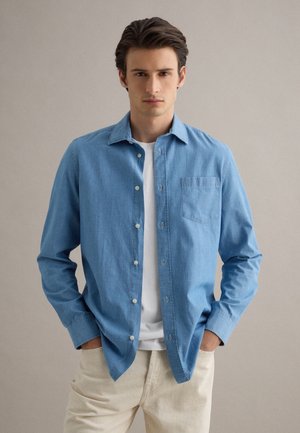 Joven con cabello corto y oscuro que lleva una camisa azul de mezclilla sin abotonar sobre una camiseta blanca y pantalones beige claro, con las manos en los bolsillos.