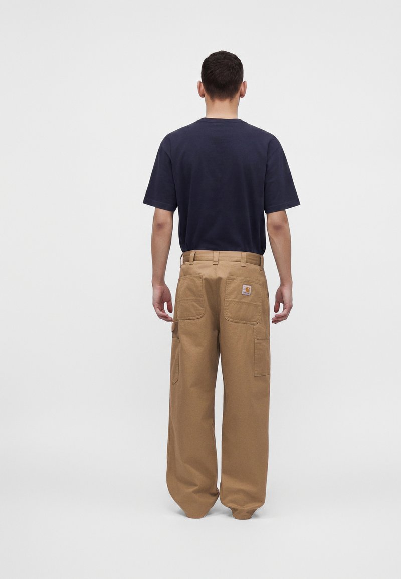 Homme portant une chemise noire à manches courtes et un pantalon de travail marron ample, debout de dos sur un fond uni.