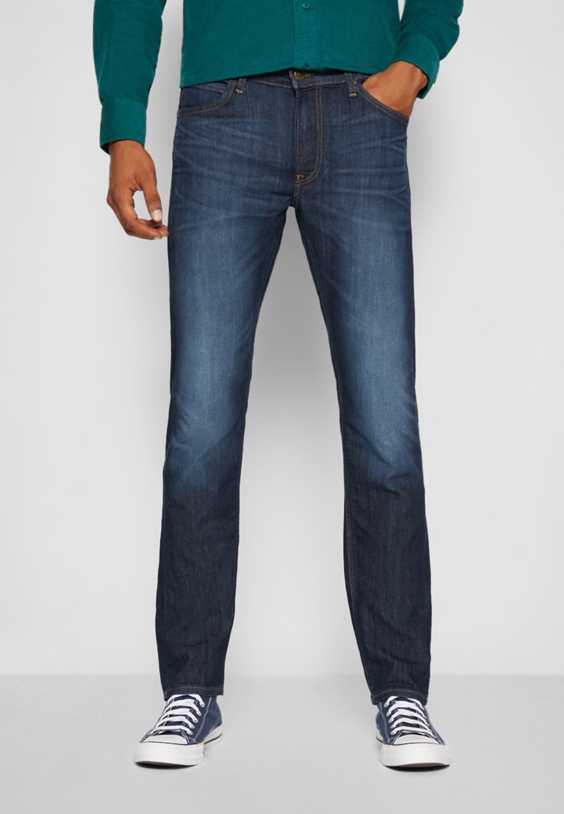 Jean en denim bleu foncé, coupe slim, lavage moyen, cinq poches et surpiqûres contrastantes, associés à des baskets blanches.