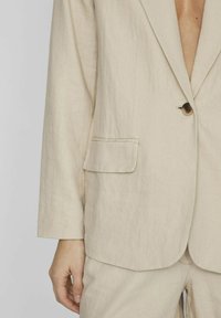 Beige linnen blazer met een enkele knoopsluiting, notch-revers en een borstzak. De stof toont een subtiele textuur en valt losjes.