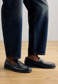 Schwarze Lederlofer mit gestickten Details und strukturierten Akzenten, getragen mit dunklem Denim, der ein lässiges und schlankes Design zur Schau stellt.