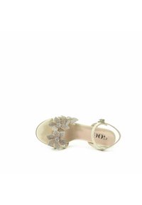 Beżowe sandały na wysokim obcasie z trzema ozdobami w kształcie kwiatów z klejnotami na pasku, z cienkim paskiem wokół kostki i gładką, błyszczącą powierzchnią.