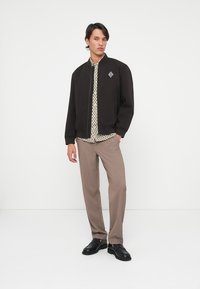 Mustan värvi bomber-jakk, millel on soonikkoes krae, allpool mustriline hele särk, heledast pruunist riidest püksid ja mustad kingad.