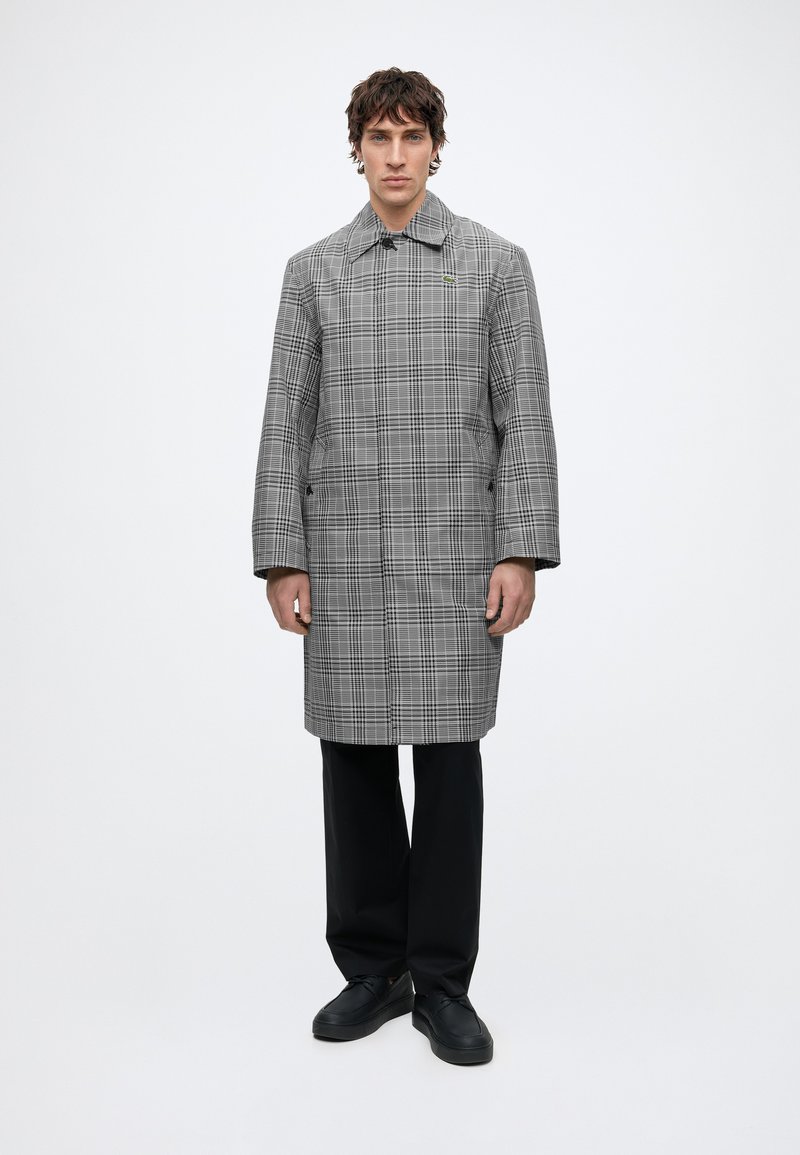 Homme debout portant un long manteau à carreaux gris, un pantalon noir et des chaussures noires sur un fond blanc uni.