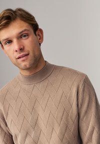 Beige Pullover mit strukturiertem Rautenmuster, geripptem Kragen und langen Ärmeln. Weicher Stoff, taillierte Passform und dezente Farbe.