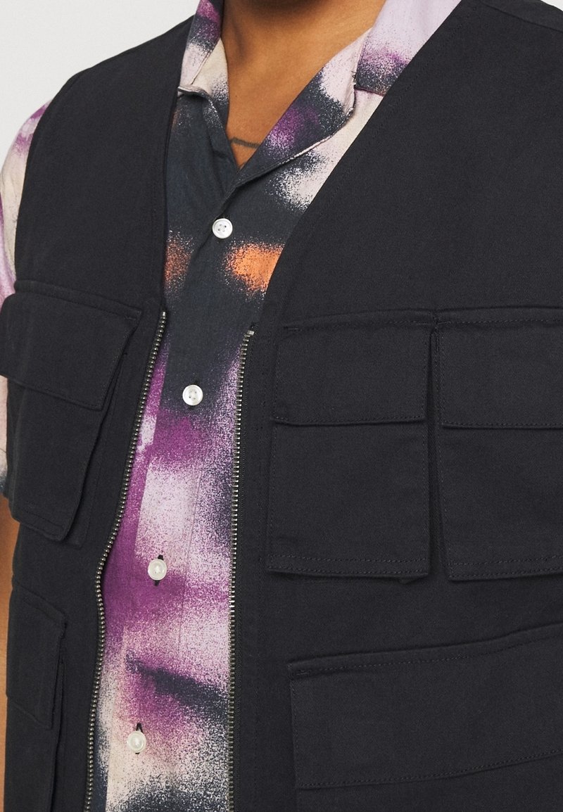 Czarna kamizelka na zamek z czterema dużymi przednimi kieszeniami, nałożona na wielokolorową, abstrakcyjną koszulę z kołnierzykiem w fioletowo-pomarańczowo-czarny wzór.