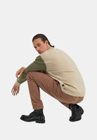 Maglione lavorato a maglia con corpo beige e maniche verdi, abbinato a pantaloni marroni. Stivali neri alla caviglia con una cintura e suola texturizzata.