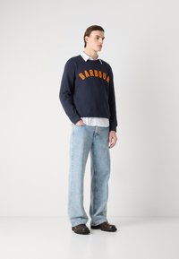 Sweatshirt azul-marinho com texto laranja "BARBOUR", combinado com jeans de perna larga azul-claro e botas castanhas escuras, estilizado sobre uma camisa branca com colarinho.