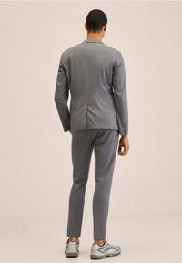 Man in een strakke grijze pak met bijpassende broek en witte sneakers, staande met zijn rug naar de kijker tegen een vlakke achtergrond.