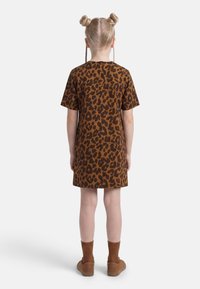 Παιδί με ξανθά μαλλιά σε δύο κότσους και πλεξούδες, φοράει καφέ φόρεμα με λεοπάρ σχέδιο, καφέ κάλτσες και καφέ παπούτσια, στέκεται γυρισμένο πλάτη.