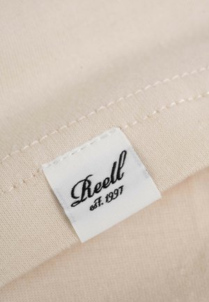 Cream-colored fabric with a textured surface, featuring a white label that reads "Reell est. 1997" in elegant black cursive.