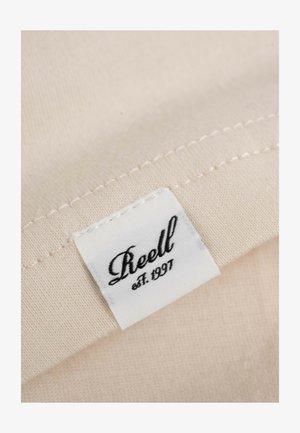 Beige Stoff mit strukturierter Oberfläche, versehen mit einem weißen Etikett, auf dem "Reell est. 1997" in eleganter schwarzer Schreibschrift steht.