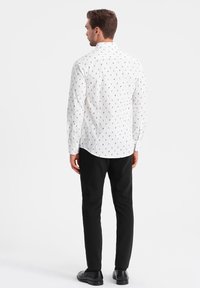 Camisa blanca con un patrón negro, de manga larga, con cuello tipo polo, combinada con pantalones negros y zapatos oscuros, vista desde la parte de atrás.