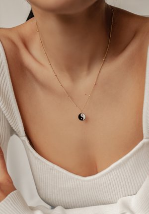 Gold chain necklace with a yin-yang pendant. The pendant features a black and white design, complementing a ribbed white top.
