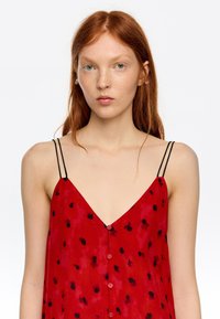 Młoda kobieta o rudych włosach i piegach, ubrana w czerwoną sukienkę bez rękawów z czarnymi kropkami i cienkimi podwójnymi ramiączkami, zwrócona przodem.