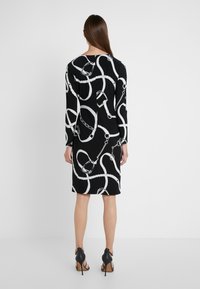 Svart klänning med ett vitt kedje- och öglmönster, långa ärmar och knälång längd, kombinerad med svarta högklackade skor.