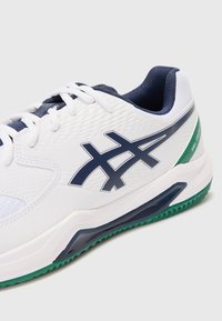 Weißer Sportschuh mit strukturiertem Obermaterial, marineblauen Akzenten und grüner Gummisohle. Verfügt über ein auffälliges Logo an der Seite. Die Schnürsenkel sind weiß.